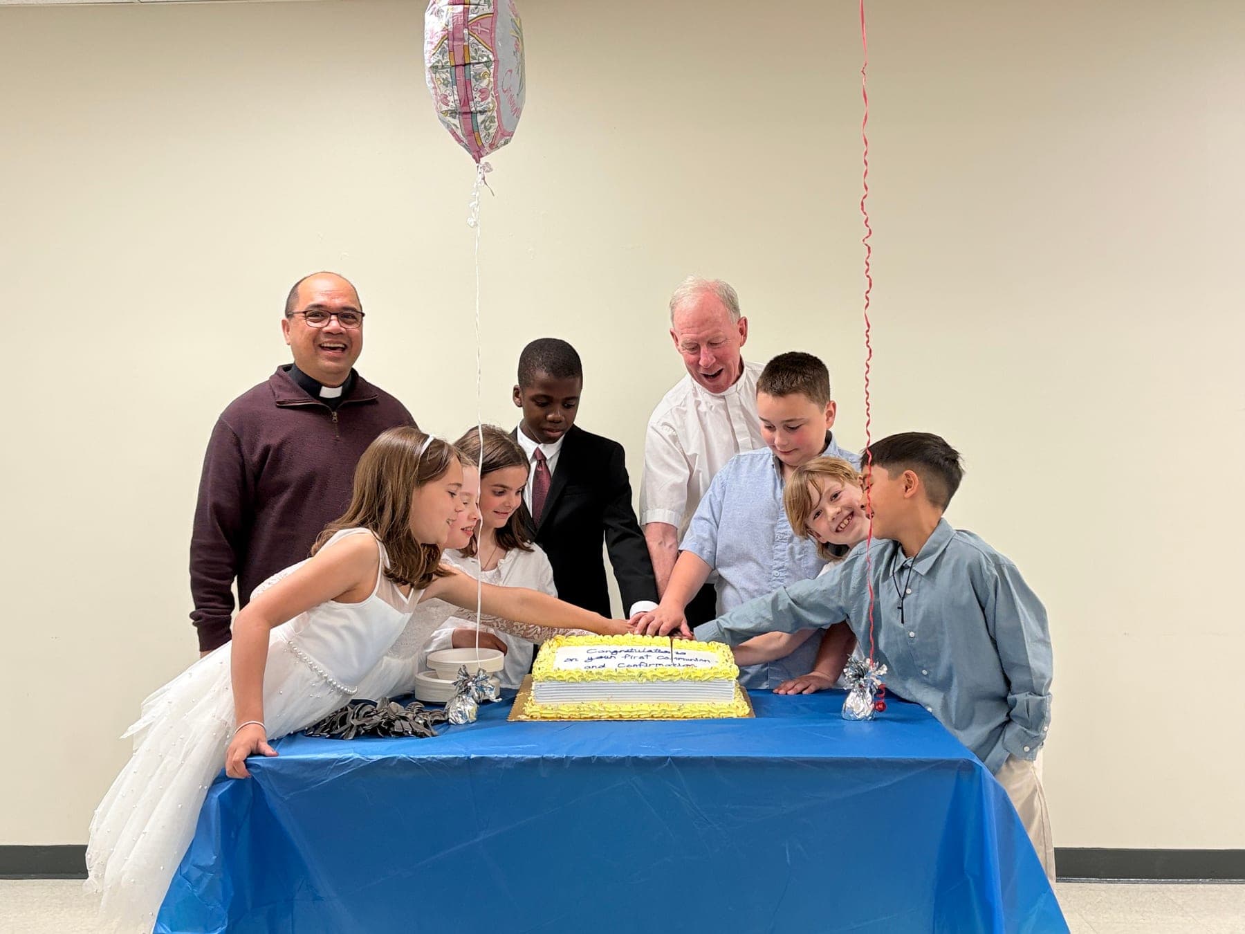 Children gathered with clergy to cut a cake after First Communion and Confirmation