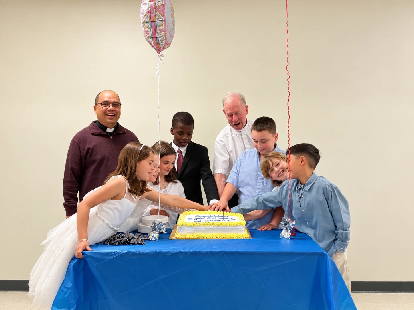 Children gathered with clergy to cut a cake after First Communion and Confirmation